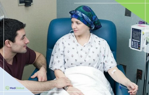 Хіміотерапія при онкології грудей Жінка в лікарняній палаті проходить хіміотерапію: сидить у кріслі з крапельницею, на голові хустка, поруч чоловік підтримує її, тримаючи за руку