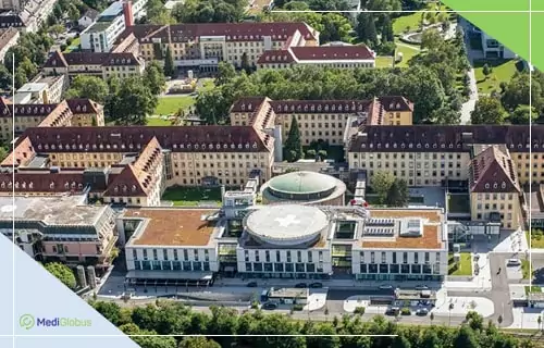 лечение лейкоза в Германии Аэрофотоснимок крупного медицинского комплекса с несколькими симметричными корпусами и центральным круглым зданием с куполом; вокруг зеленые деревья, дороги и автобусы у входа.