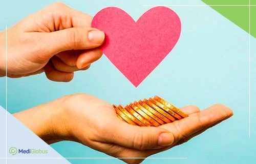 Благодійні фонди допомагають збирати кошти на лікування руки людини у крупному плані: в одній руці — вирізане з червоного паперу серце, в іншій — золоті монети.
