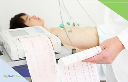 Как предотвратить болезни сердца Пациент проходит электрокардиографическое обследование: лежит на кушетке с подключёнными электродами на груди, врач держит в руках распечатку ЭКГ с графиком сердечных сокращений; рядом стоит аппарат, фиксирующий данные.