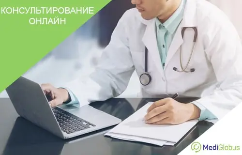 Онлайн-консультация с доктором Врач в белом халате и со стетоскопом вокруг шеи сидит за столом и работает с ноутбуком и документами