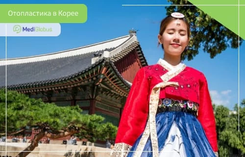 Коррекция ушей - цена в Корее Женщина в традиционном корейском наряде стоит на фоне старинного дворца с изогнутой крышей и декоративными элементами