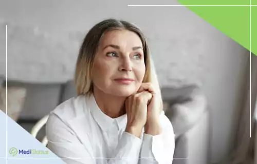 Жінка середнього віку в білій сорочці задумливо дивиться вбік, сидячи у світлій кімнаті