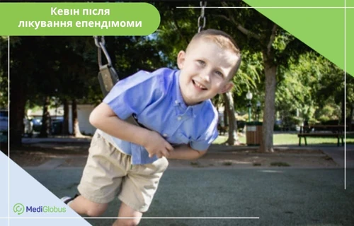 Відгуки про лікування епендимоми Маленький хлопчик зі світлим волоссям у синій сорочці та бежевих шортах, який усміхається та катається на гойдалці на дитячому майданчику в парку; зліва надпис - "Кевін після лікування епендімоми"